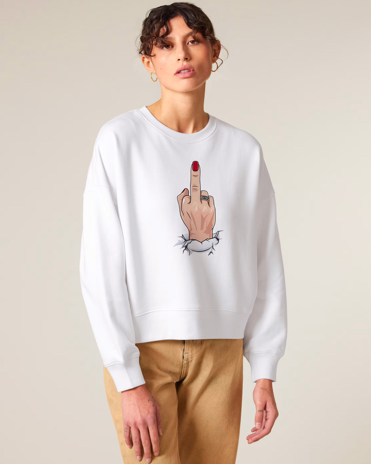 A woman wearing tan pants and a white sweatshirt featuring an illustration of a hand with red nails making a middle finger gesture.