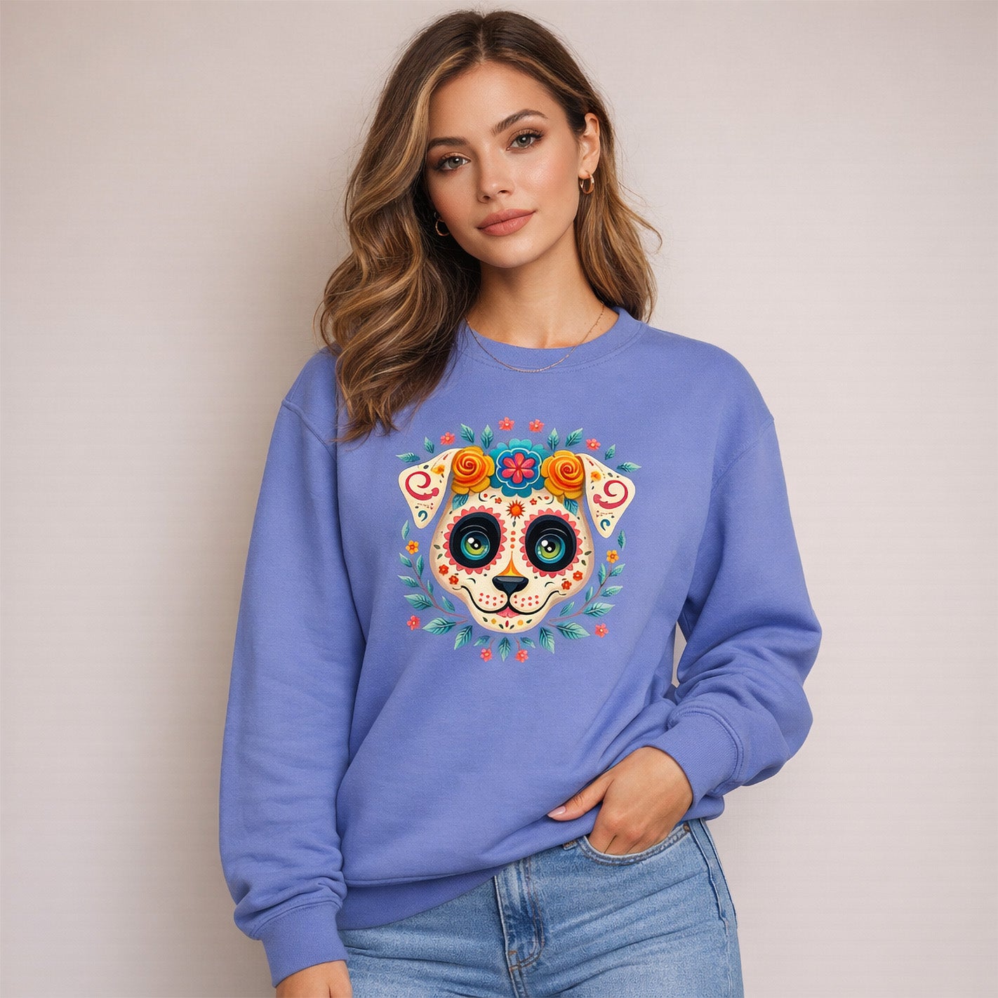 A woman in a blue sweatshirt featuring a colorful, decorative dog face design inspired by Day of the Dead motifs stands against a plain, light-colored background.