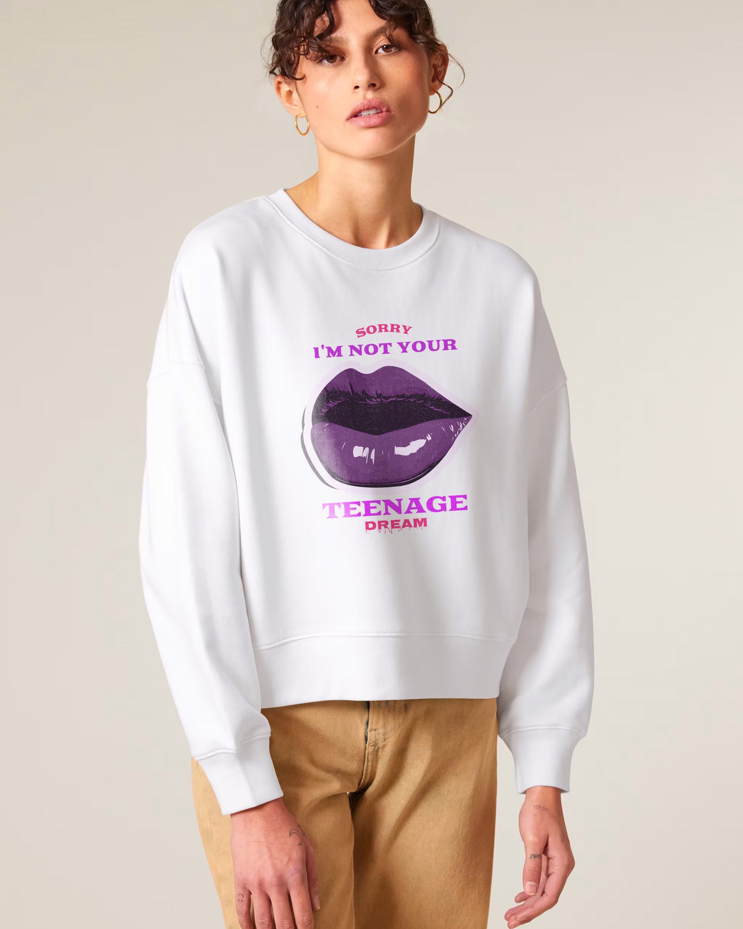 A woman wearing a white sweatshirt with a large graphic of purple lips and the text "SORRY I'M NOT YOUR TEENAGE DREAM" in pink and purple letters, paired with tan pants.