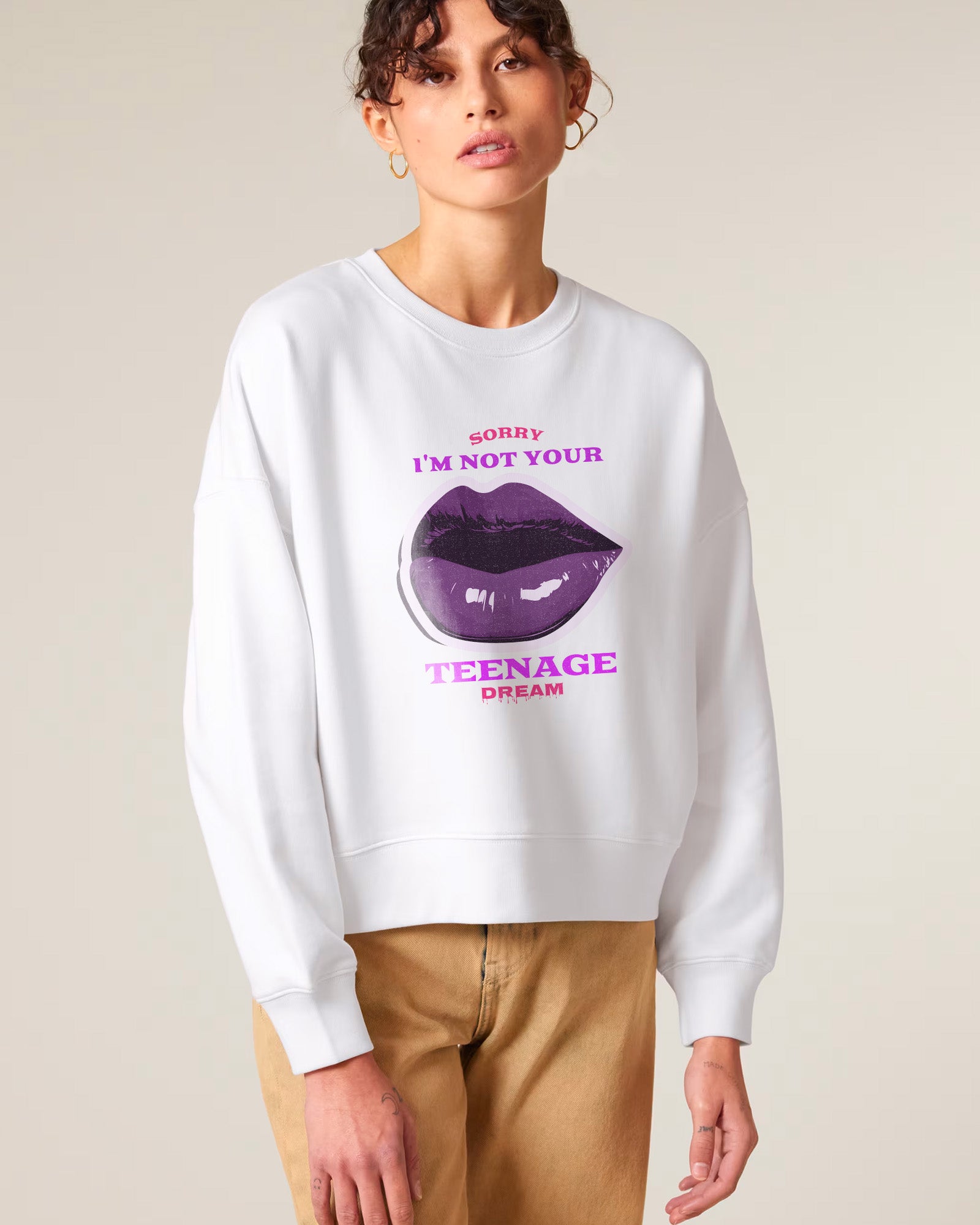 A woman wearing a white sweatshirt with a large graphic of purple lips and the text "SORRY I'M NOT YOUR TEENAGE DREAM" in pink and purple letters, paired with tan pants.