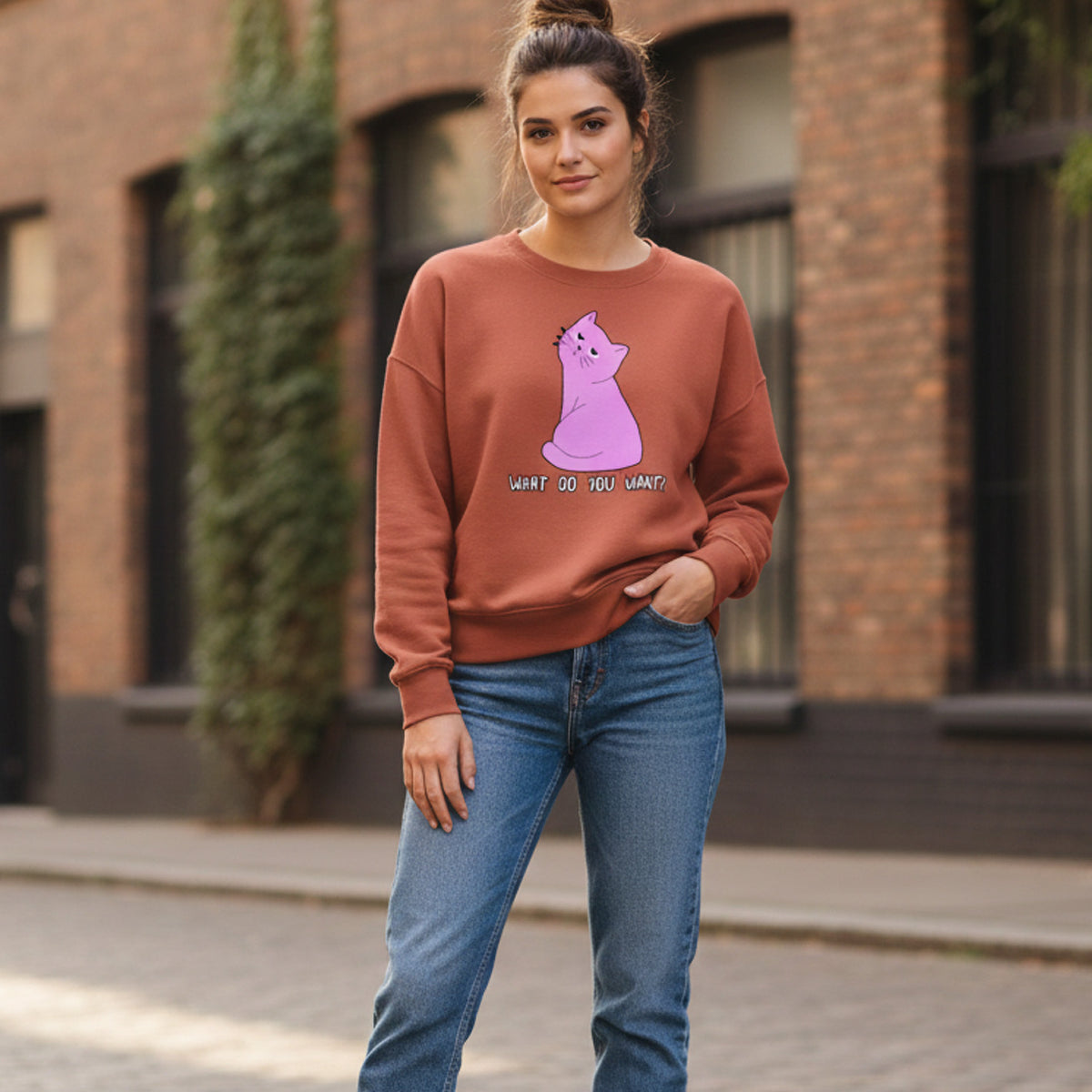 A young woman stands on a cobblestone street wearing jeans and a rust-colored sweatshirt featuring a pink cartoon cat and the text "WHAT DO YOU WANT?" The background shows a brick building and some greenery.