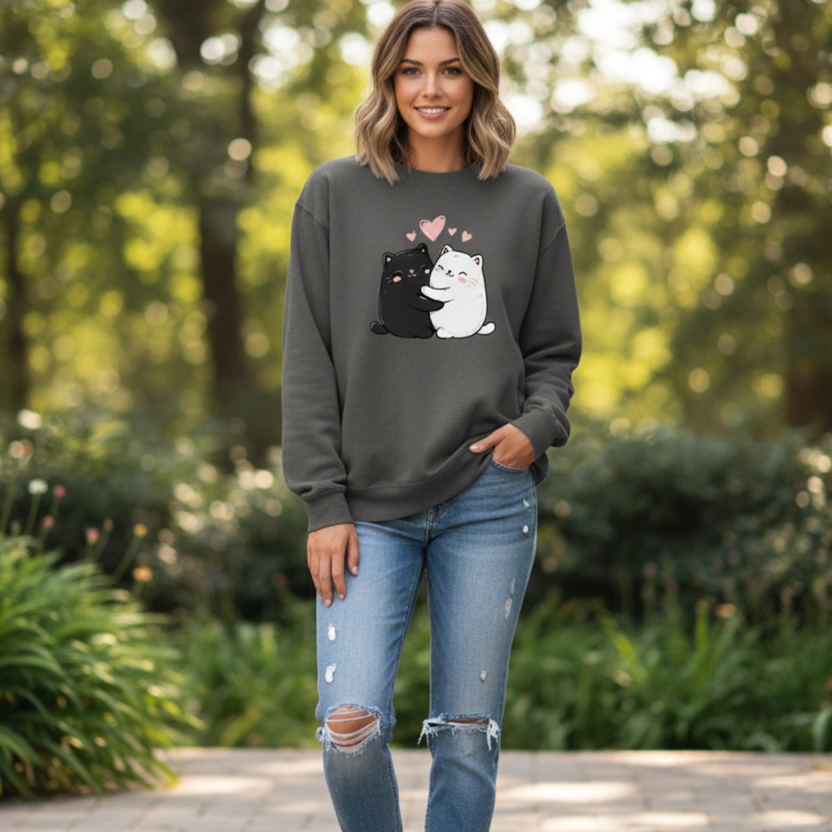 A woman stands outdoors in a garden, smiling and wearing a dark sweatshirt with a cute graphic of a black and a white cat with pink hearts above them. She is also wearing ripped blue jeans.