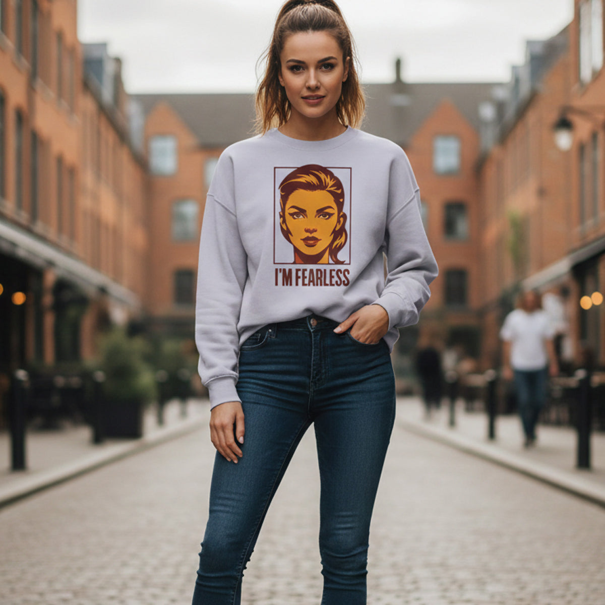 A woman stands confidently on a cobblestone street, wearing a light-colored sweatshirt with a bold graphic of a woman's face and the words "I'M FEARLESS," and dark jeans. The background features brick buildings.