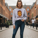 A woman stands confidently on a cobblestone street, wearing a light-colored sweatshirt with a bold graphic of a woman's face and the words "I'M FEARLESS," and dark jeans. The background features brick buildings.