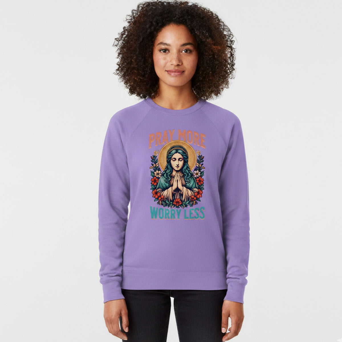 A woman wearing a lavender sweatshirt with an illustration of a praying figure surrounded by flowers and the text "Pray More Worry Less" stands facing forward, smiling slightly against a plain background.