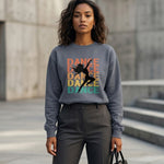A woman stands outdoors wearing a gray sweatshirt with the word "DANCE" in colorful letters and a silhouette of a dancer. She has wavy hair, gray trousers, and holds a black bag. The background features concrete walls and steps.