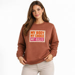 A woman wearing a brown sweatshirt with the bold text "MY BODY MY CHOICE MY RULES" in orange, yellow, and pink stands against a plain white background.