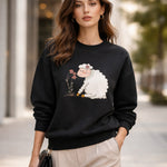 A woman stands outdoors wearing a black sweatshirt with a cute sheep graphic eating flowers. She has loose brown hair, gold hoop earrings, and holds a small black purse. The background is a city street with blurred trees and buildings.