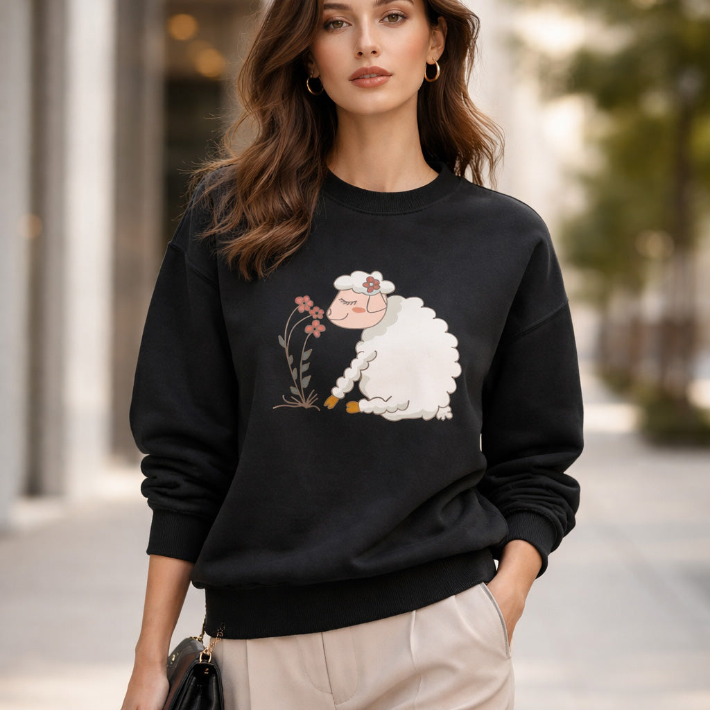 A woman stands outdoors wearing a black sweatshirt with a cute sheep graphic eating flowers. She has loose brown hair, gold hoop earrings, and holds a small black purse. The background is a city street with blurred trees and buildings.