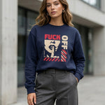 A woman standing outdoors wears a navy sweatshirt with bold text reading "FUCK OFF" and an abstract graphic design. She has wavy hair and is dressed in gray trousers, posing confidently on a city street.