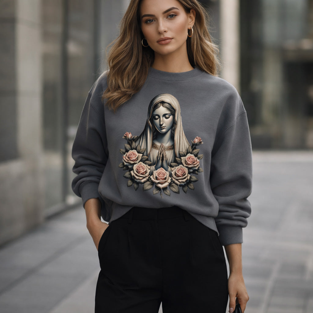 A woman stands outdoors wearing a gray sweatshirt featuring an artistic image of the Virgin Mary surrounded by roses. She has loose, wavy hair and is dressed in black pants, with a modern urban background behind her.