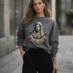 A woman stands outdoors wearing a gray sweatshirt featuring an artistic image of the Virgin Mary surrounded by roses. She has loose, wavy hair and is dressed in black pants, with a modern urban background behind her.