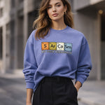 A woman wearing a blue sweatshirt with a periodic table design spelling "Sarcasm" and the phrase "May occur periodically," standing outdoors in an urban setting.