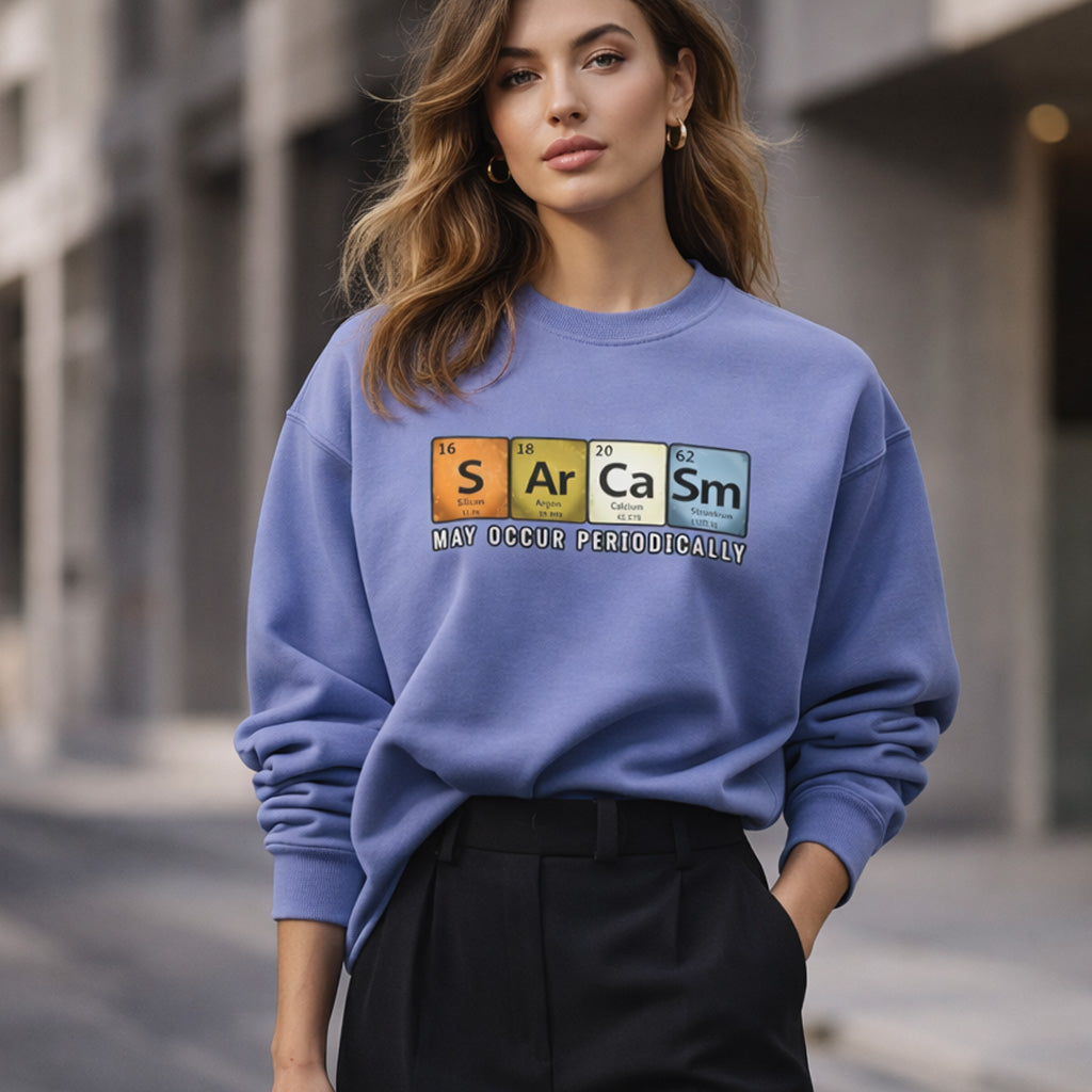 A woman wearing a blue sweatshirt with a periodic table design spelling "Sarcasm" and the phrase "May occur periodically," standing outdoors in an urban setting.