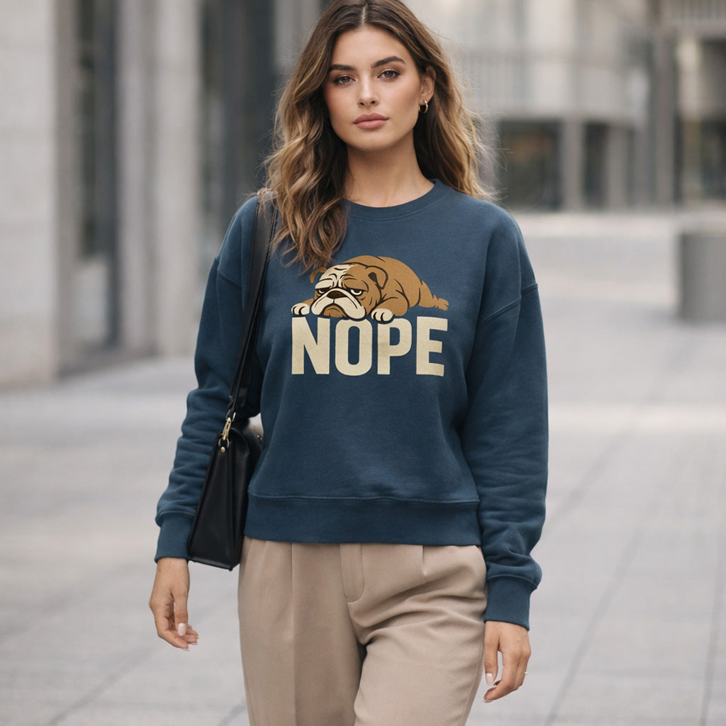 A woman with wavy hair walks outside wearing a blue sweatshirt featuring a cartoon bulldog lying down above the word "NOPE," and beige pants. She carries a black shoulder bag and appears confident.