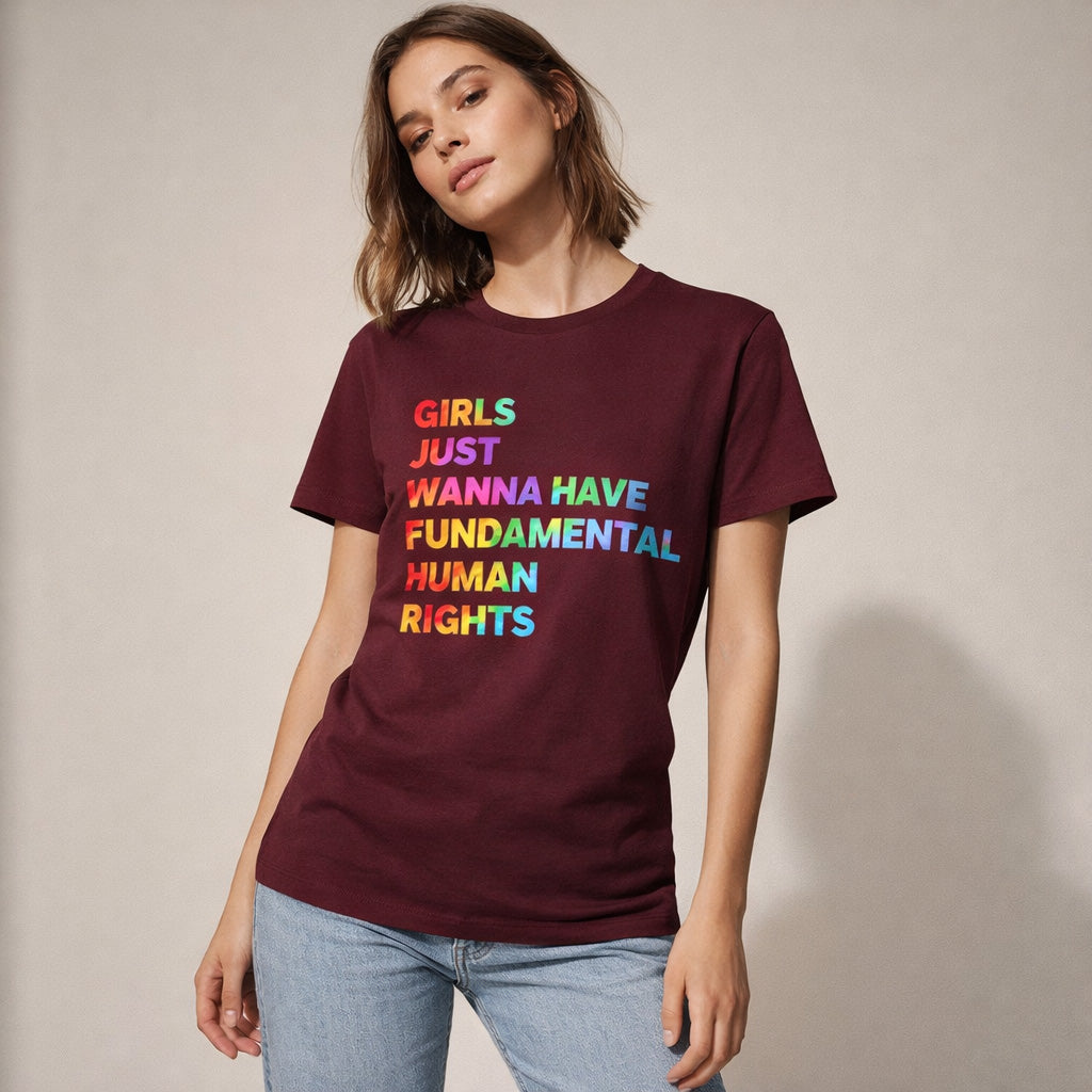 A woman in a maroon t-shirt with colorful text reading "Girls just wanna have fundamental human rights" stands against a plain background, wearing light blue jeans and looking confidently at the camera.