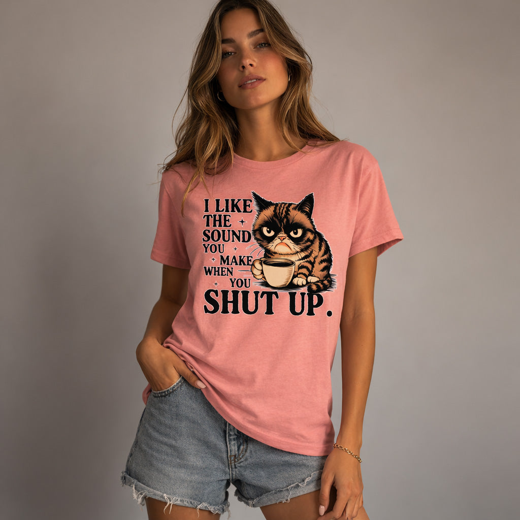 A woman in a pink t-shirt and denim shorts stands with one hand in her pocket. The shirt features a grumpy cat holding a coffee cup and the text: "I like the sound you make when you shut up.