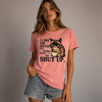 A woman in a pink t-shirt and denim shorts stands with one hand in her pocket. The shirt features a grumpy cat holding a coffee cup and the text: "I like the sound you make when you shut up.