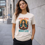 A woman stands outdoors wearing a t-shirt with an illustration of a praying figure surrounded by flowers and the words "PRAY MORE WORRY LESS" printed above and below the image.