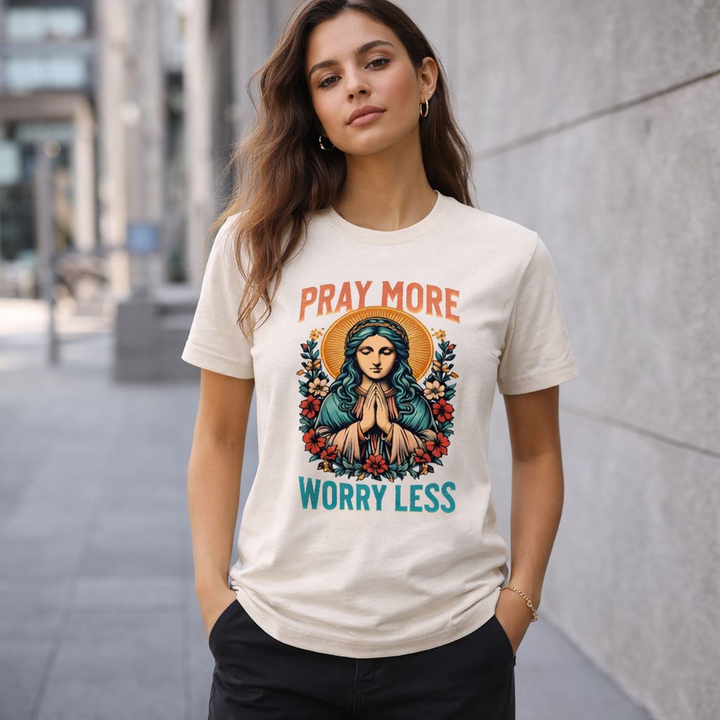 A woman stands outdoors wearing a t-shirt with an illustration of a praying figure surrounded by flowers and the words "PRAY MORE WORRY LESS" printed above and below the image.