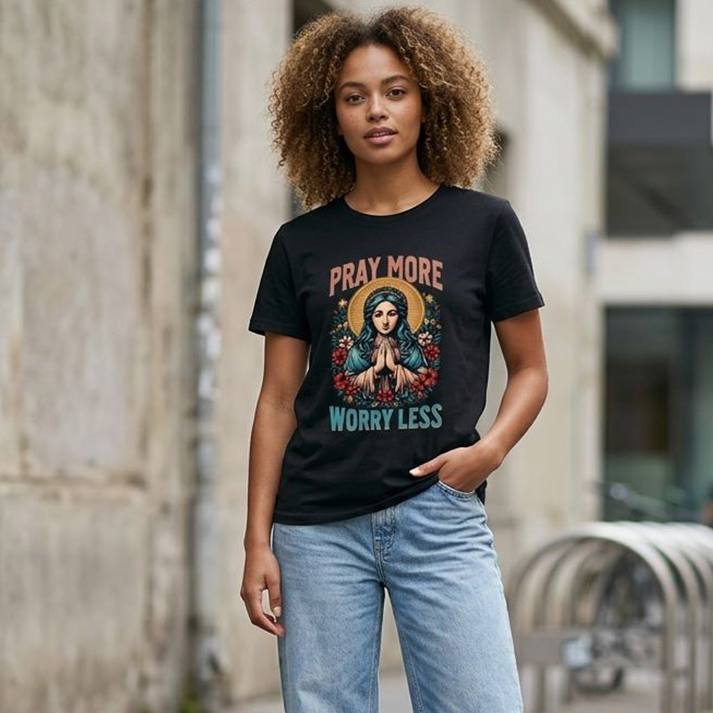 A woman with curly hair stands outdoors wearing a black T-shirt that reads "Pray More Worry Less" with an illustration of a praying woman surrounded by flowers. She has one hand in her pocket and stands near a bike rack.