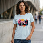 A woman stands outdoors wearing a light blue t-shirt with the word "WOMAN" in bold, colorful, graffiti-style letters. The background shows a modern city street with buildings and a sidewalk.
