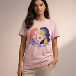 A woman in a light pink T-shirt with an illustration of two women facing each other, a heart between them, and the text "beauty of love." She stands with one hand in her pocket against a neutral background.