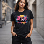 A woman standing on a city street wears a black t-shirt with colorful graffiti-style text reading "WOMAN." She has long hair, hands in pockets, and looks confidently at the camera.