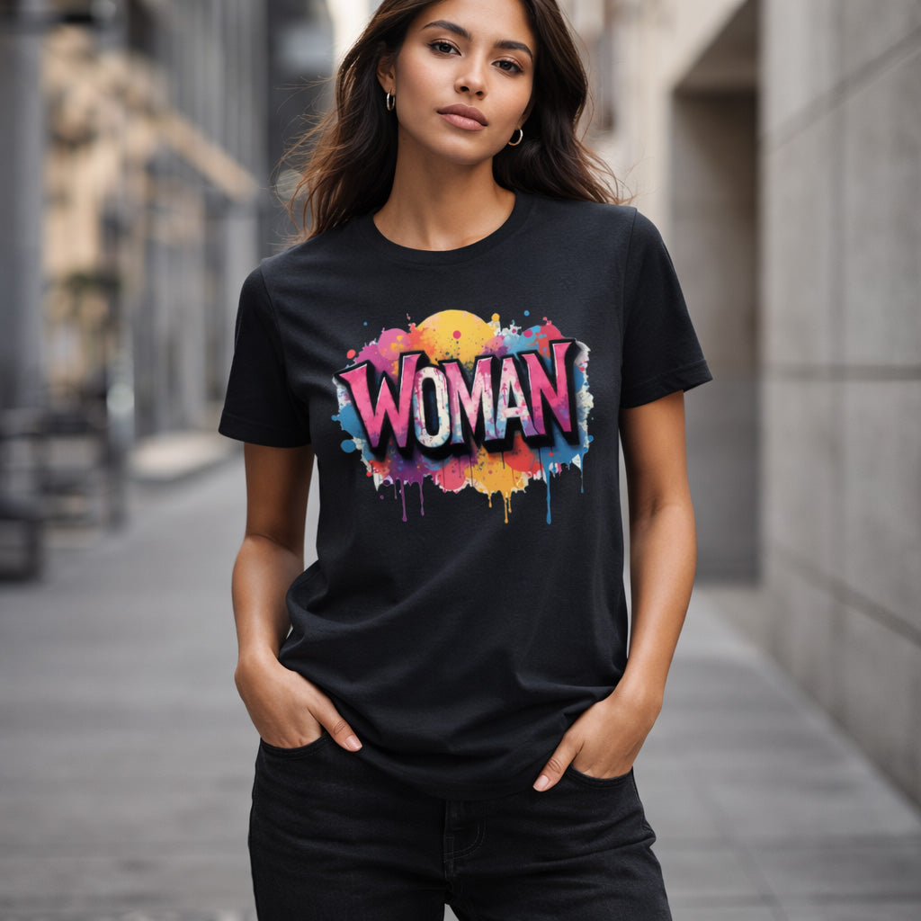 A woman standing on a city street wears a black t-shirt with colorful graffiti-style text reading "WOMAN." She has long hair, hands in pockets, and looks confidently at the camera.