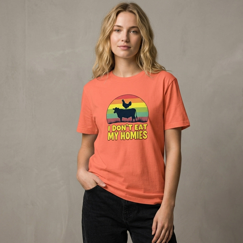 A woman wearing a coral t-shirt with a retro-style graphic of a cow and chicken, and the words "I DON'T EAT MY HOMIES," stands against a neutral background with one hand in her pocket.