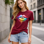 A woman wearing a burgundy T-shirt with a colorful, dripping rainbow lips graphic stands on a city street. She has long blonde hair and is also wearing light denim shorts. The background features buildings and a sidewalk.