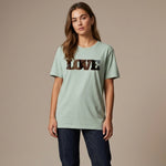A woman with long brown hair wears a light green T-shirt with the word "LOVE" in bold, dark letters on the front. She is standing against a plain beige background and has a neutral expression.