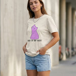 Woman wearing a white t-shirt with a purple cat graphic and text, standing against a light-colored wall.