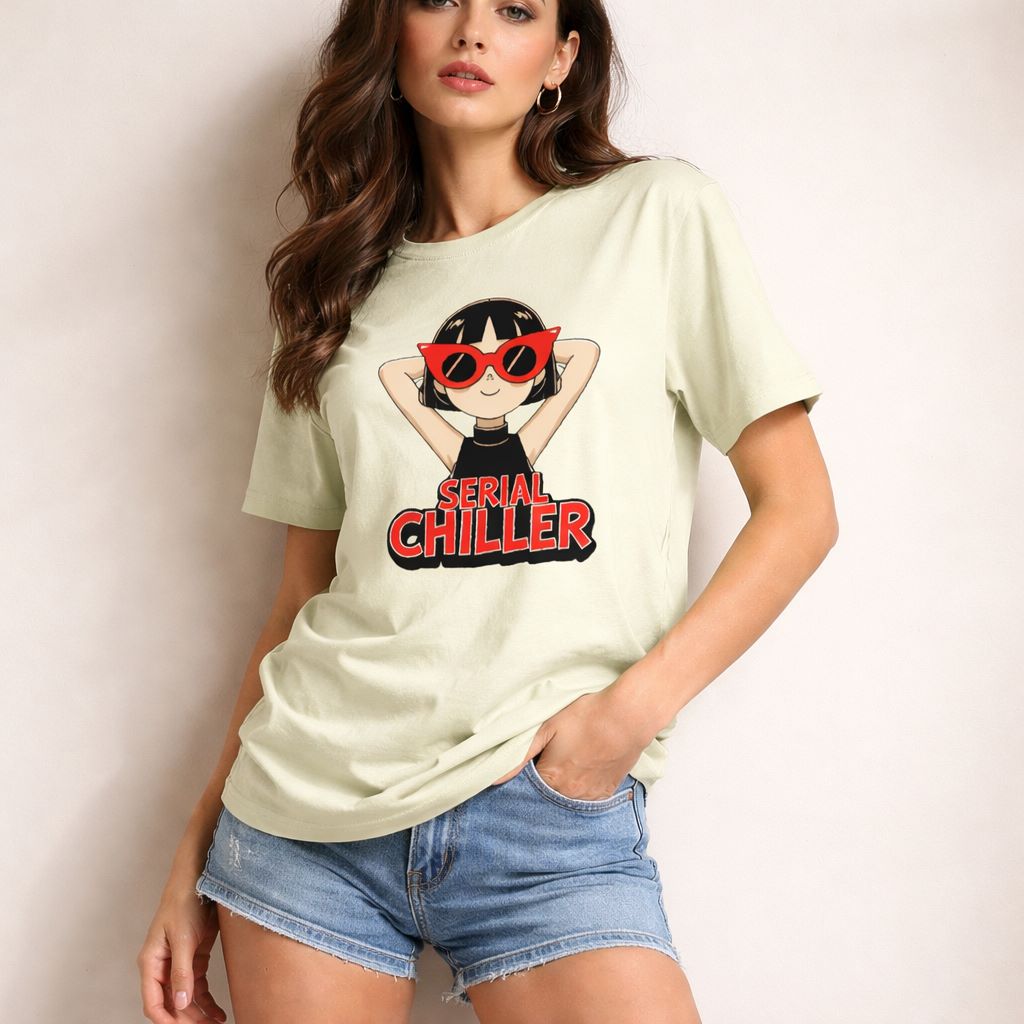 A woman wearing denim shorts and a light-colored T-shirt with a cartoon girl in red sunglasses and the words "Serial Chiller" printed on it stands against a plain background, holding part of her shirt.